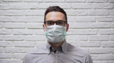 Kareli gri gömlekli yakışıklı beyaz erkek esmer koruyucu turkuaz bir tıbbi maske takıyor ve beyaz tuğla duvar arkasında dişleriyle gülümsüyor. Maske takılır ve bir kulağa tutunur..