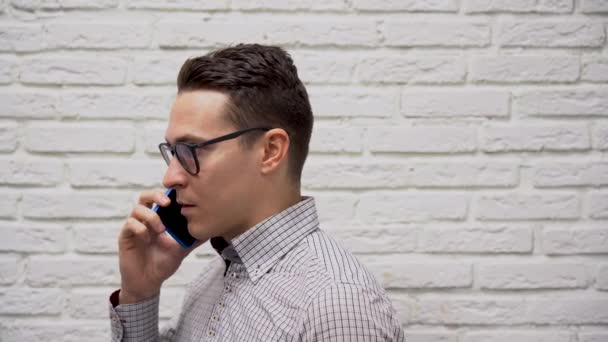 身穿格子灰色衬衫 戴着黑色眼镜 拿着蓝色手机在白色砖墙背景下与客户交谈的年轻黑人客户经理 商人手里拿着电话 面带微笑 — 图库视频影像