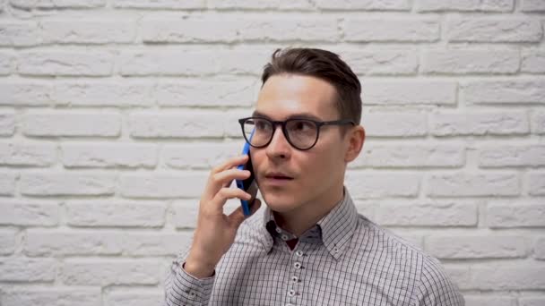 严肃的白人商人 戴着黑色眼镜 身穿格子灰色衬衫 拿着蓝色电话交谈 他同意了 结束电话 看着白色砖墙背景上的电话 — 图库视频影像