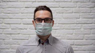 Siyah gözlüklü beyaz tenli genç adam, yüzünde koruyucu tıbbi maske olan kareli bir gömlek, gülücükler ve dans, beyaz tuğlalı arka planda kazanan jest. İnsanoğlu virüse karşı zaferle sevinir.