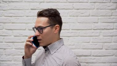 Siyah gözlüklü, kareli gri gömlekli genç bir esmer muhasebe müdürü müşterisiyle mavi bir telefonla beyaz tuğladan bir arka planda konuşuyor. İş adamı telefonu elinde tutuyor ve gülümsüyor..