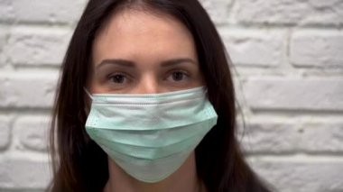 Uzun saçlı, kahverengi gözlü, koruyucu tıbbi maskeli, güzel esmer bir kız başını sallıyor ve beyaz tuğlalı bir arka plana hayır diyor. İnkar jesti.