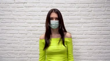 Yüzünde koruyucu tıbbi maske takan yeşil ceketli tatlı, genç esmer kadın beyaz tuğla duvar arkasında bir korku hissediyor. Corona virüsünden korkuyor. Korku kalbi yakalar..