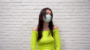 Yeşil ceketli güzel esmer kadın, beyaz tuğla duvarlı arka planda Corona virüsü yüzünden koruyucu maske takıyor. Mutlu bir kız gülümser, dans eder, neşelenir ve kameraya bakar.