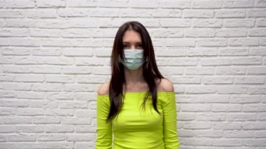 Yüzünde koruyucu tıbbi maske olan yeşil ceketli, yakışıklı beyaz esmer kadın gözlerini beyaz tuğla duvara doğru açıyor. Corona virüsünden korkuyor. Korkuyu hisseder.