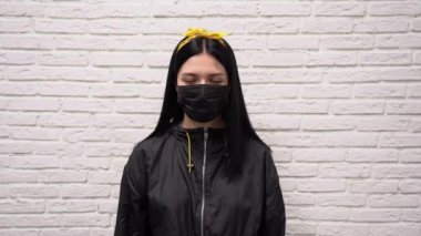Siyah ceketli, yüzünde koruyucu bir maske takmış beyaz tuğla duvardaki virüsten dolayı yüzünde uzun saçlı beyaz bir kız. Kadın kameraya bakar, güler ve gülümser..
