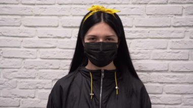 Sarı bantlı, uzun siyah saçlı beyaz kız yüzüne koruyucu tıbbi maske takıyor. Kız şaşırdı ve beyaz tuğlalı duvara karşı şaşkınlık içinde kaldı..
