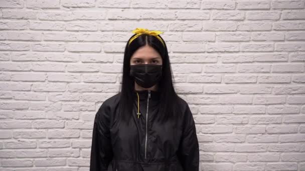保護医療マスクで黒のジャケットに黄色のヘッドバンドと美しい若い白人女性は彼女の頭を振る と言ういいえ白いレンガの壁の背景に近い 否定のジェスチャー — ストック動画