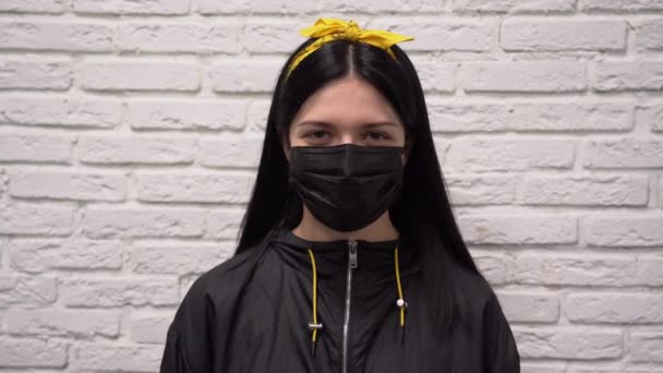 黄色のヘッドバンドに長い黒髪の若いかわいい白人の女の子は 顔に保護医療マスクを着用してください 女の子驚きと混乱の中で白いレンガの壁の背景に対して閉じるアップ — ストック動画