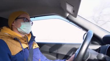 Kışın sarı şapkalı, yüzünde siyah gözlüklü, virüsten koruyucu bir maske takan, sıcak mavi ceketli, çekici beyaz bir adam parlak güneşli bir günde araba kullanıyor. İç görünüm.