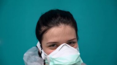 Beyaz koruyucu tulumlu, yüzünde solunum cihazı ve sağlık maskesi olan beyaz, beyaz, beyaz, beyaz bir kadın gözlük, fermuar ceket takıyor ve mavi arka planda kapüşon takıyor. Kız gülümsemedir. 
