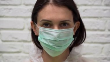 Yeşil koruyucu takım elbiseli, kahverengi saçlı güzel beyaz kadın yüzüne solunum cihazı ve tıbbi maske takıyor ve beyaz tuğlalı arka planda kayboluyor. Kız gülümsüyor.