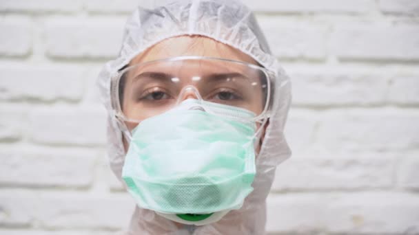 白い保護スーツの医療女性はマスク マスク マスクを着用し 恐怖感を感じます 恐怖と恐怖のCovid 19ウイルス 目を大きく開けて レンガの背景を見ると — ストック動画