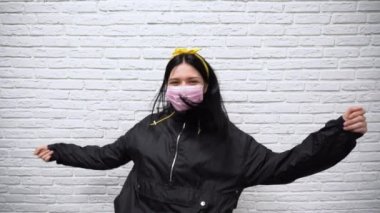 Uzun siyah saçlı, spor ceketli ve pembe koruyucu maskeli sarı bantlı güzel Rus kadın beyaz tuğla duvarın arka planında ağır çekimde dans ediyor..