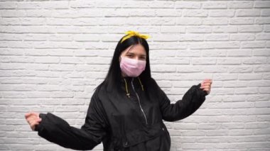 Beyaz tuğlalı bir duvarın arka planında uzun siyah saçlı, sarı bantlı, tek kullanımlık pembe filtre maskeli, çekici, mutlu bir Rus kızı ağır çekimde dans ediyor..
