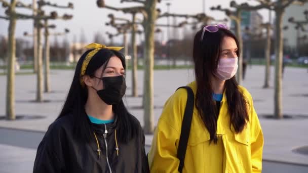 两名身穿黑色和黄色夹克 戴着医疗一次性防护面具的漂亮的意大利黑发女孩在日落时分走在城市街道上 女性朋友快乐 说话慢动作 — 图库视频影像