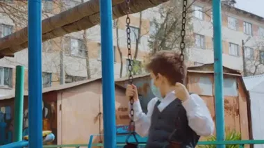 Okul kıyafetli, kısa sarı saçlı, turkuaz koruyucu tıbbi maskeli bir çocuk güneşli bir günde paslı metal zincirlerle salıncakta sallanıyor. Covid 19 koruma kavramı