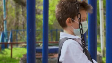 Yalnız küçük öğrenci koruyucu bir maske takıyor, okul kıyafeti giyiyor ve başını eğiyor. Sarışın çocuk üzgün, metal zincirle salıncakta sallanıyor ve bulanık yeşil arka planda yere bakıyor..