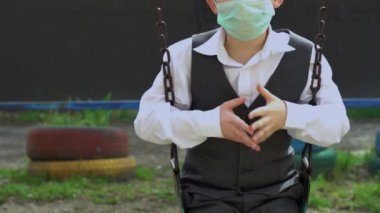 Beyaz gömlekli, gri yelekli, turkuaz koruyucu maskeli, yüzünde salıncakta sallanan yalnız bir çocuk. Gözbebeği şans diliyor, yakın çekim. Koronavirüs tehdidi nedeniyle karantina süresi.