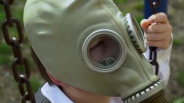 Beyaz okul tişörtlü yalnız bir çocuk, oyun parkında, paslı metal zincirli, askeri koruyucu gaz maskeli bir salıncakta oturuyor. Sıkılmış çocuk insanlardan izole edilmiş güneşli günlerde karantinaya alınır.. 