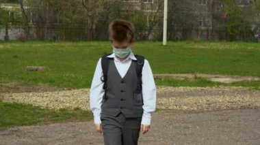 Okul çantalı, başı eğik yalnız bir öğrenci turkuaz koruyucu tıbbi maske takıyor, yere bakıyor, güneşli bir günde yeşil çimen tarlasının arka planında üzgün bir şekilde yürüyor.