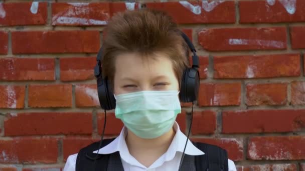 阳光明媚的一天 身穿校服 戴着防护医疗面罩的白人帅哥 在红砖墙的背景下 用黑色大耳机听音乐 学生在病毒感染期间关闭 — 图库视频影像
