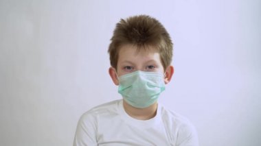 Beyaz arka planda izole edilmiş yeşil koruyucu bir tıbbi maske takan uzun kollu, kısa saçlı, beyaz saçlı beyaz bir çocuk. Çocuğun burnu kaşınıyor ve kaşınıyor. Kaşınıyor..