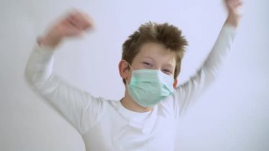 Uzun kollu güzel sarışın adam, beyaz arka planda Corona virüsünden koruyucu bir maske takıyor. Küçük mutlu bir çocuk gülümser, dans eder, neşelenir ve kameraya bakar..