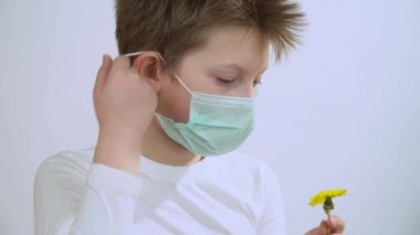 Küçük çocuk elinde sarı bir karahindiba tutuyor, bak, koruyucu tıbbi maskeyi çıkar, çiçeği kokla, nefes al ve koku al. Yakışıklı çocuk beyaz arka plana maske takmış..