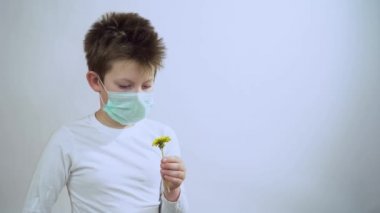 Kısa sarı saçlı küçük çocuk elinde sarı bir karahindiba tutuyor, bak, koruyucu tıbbi maskeyi çıkar, çiçek kokla, aroma solu. Adam yüzüne beyaz arka planda bir maske takmış. Yakın plan..