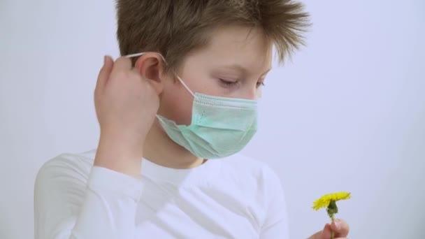 小男孩手里拿着一朵黄色的蒲公英 摘下防护的医疗面具 嗅着花 呼吸着 闻到了香味 一个英俊的孩子把他的脸戴在白色背景的面具上 — 图库视频影像