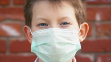 Tıbbi koruyucu maskeli gri gözlü, güzel, genç, beyaz, sarışın bir çocuk güneşli bir günde kameraya bakar ve yavaş çekimde yaklaşır. Çocuk Coronavirus covid-19 virüsünden korkmaz..