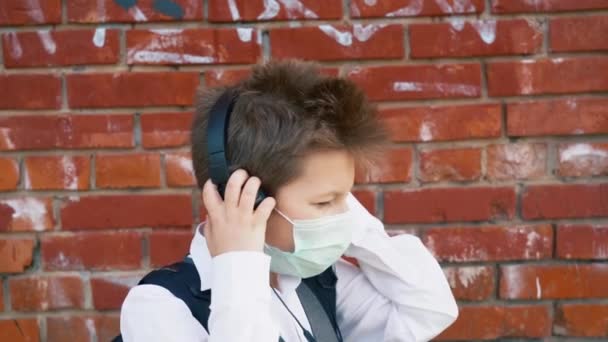 穿着校服 戴着医用防护面罩的英俊小学生 头上戴着大大的黑色耳机 在红砖墙上听音乐 动作缓慢 男孩在检疫期间享受生活 — 图库视频影像