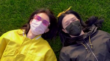 Cute Caucasian girls in black and pink medical protective faces masks lie on green grass and show the middle finger on a. Women give with hands gesture impolite and rude - fuck off. View from above.