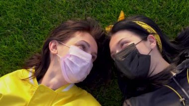 Siyah ve pembe tıbbi koruyucu yüzlü iki beyaz kız yeşil çimlerin üzerinde duran ceketli maskeler takıyor. Kadınlar birbirlerine aşkla bakar ve güneşli bir günde yakınlaşırlar. Yukarıdan bak. Coronavirus covid-19 virüsü tehdidi nedeniyle kilitlenin.