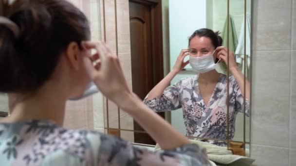 美しい白人のブルネットの女性がパジャマを着て 保護医療の顔のマスク 自宅のバスルームで鏡の前で自分自身を賞賛する Covid19ウイルス分離期間 — ストック動画