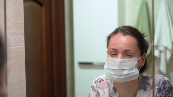 漂亮的白种人黑发女孩 头戴睡袍 戴蓝色防护面具 在镜子里看 做鬼脸 在浴室里笑 根据Coronavirus Covid 19病毒进行检疫 — 图库视频影像