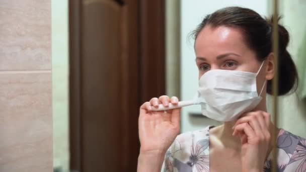隔離された素敵な白人の美しいブルネットの女の子は青い保護医療マスクを着用し バスルームの鏡の前で歯ブラシで彼女の歯を磨きます 家での典型的な隔離朝 — ストック動画
