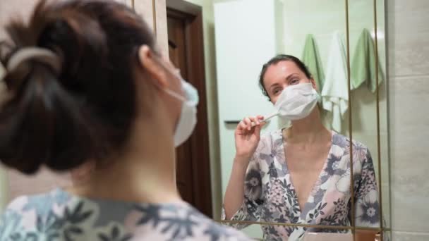 夜のパジャマで美しいブルネットの女の子は ウイルス ブラシの歯から保護医療面マスクを着用し バスルームで鏡を閉じて見てください 隔離だ 緊急事態だ パンデミック時の隔離 — ストック動画