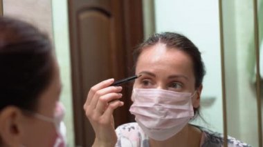 Gece makyajlı, koruyucu pembe maskeli beyaz bir kadın banyoda aynanın önünde siyah bir kalemle kaşlarını boyuyor. Evde yalnızlık.