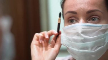 Mavi koruyucu tıbbi yüz maskesi takmış güzel beyaz bir kadın kaşlarını fırçayla düzeltiyor ve yakından tarıyor. Kız aynaya bak. Salgın sırasında karantina. Tecrit