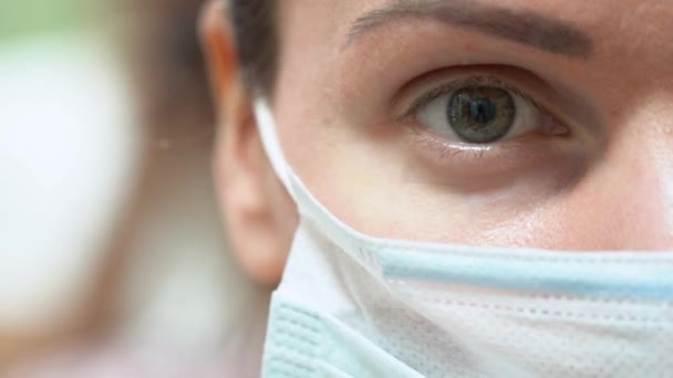 一个疲惫的绿眼睛的医生女孩盼望着模糊的背景 身穿蓝色一次性防护口罩的年轻女子的画像 宏观观点准确 病毒概念 — 图库视频影像