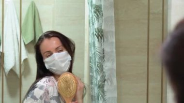 Koruyucu mavi yüzlü, çiçek pijamalı virüs maskeli, beyaz, mutlu, esmer kadın. Evdeki banyo aynasının önünde tarakla imza at. Kız tecrit döneminde hayatın tadını çıkarıyor..