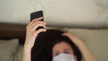 Koruyucu tıbbi maske takan bekar, güzel bir kadın bej yatakta uzanıyor ve siyah telefonun üst görüntüsünde selfie çekiyor. İyi bir karantina sabahı ve kovid19 virüs tehdidi..
