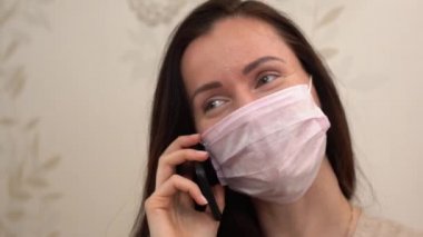 Pembe koruyucu sağlık maskeli esmer kızın portresi evde cep telefonuyla konuşuyor. Güzel bir kadın söyler, gülümser, yakından güler. İzolasyon haftasonunda, içerde iyi eğlenceler..