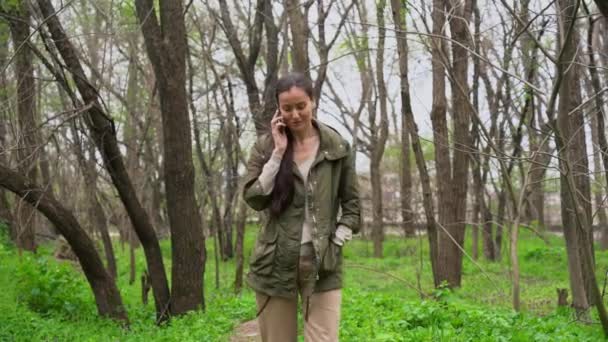 年轻美丽的白种人姑娘 棕色长发 穿着温暖的夹克 一个人走在绿草如茵的空旷的森林里 用手机交谈 在寒冷的早晨 在户外大笑 — 图库视频影像