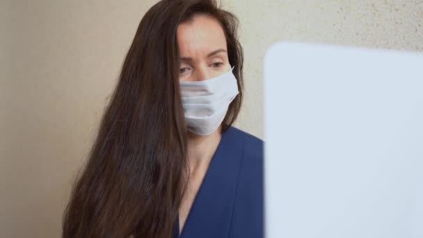 長い茶色の髪を持つ美しい女性は ノートパソコンを使用して 医療面のマスク 古典的なスーツを着て タイプのテキスト 自宅のオフィスの机の近くに電子メールで返信します 病気のビジネス女性はパンデミックの時にコンピュータで働く — ストック動画