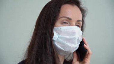 Güzel, beyaz koruyucu maskeli, gri arka planda izole bir şekilde cep telefonuyla konuşan beyaz, beyaz, beyaz bir kız. Kumandalı kahverengi saçlı genç bir iş kadını. Koronavirüsle korunma.
