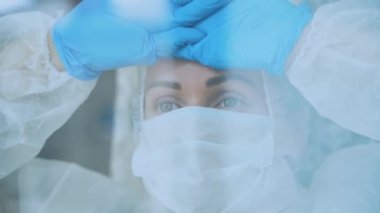 Tıbbi maske takmış beyaz koruyucu takım elbiseli, cam camına yaslanmış mavi eldivenli güzel bir doktor portresi. Üzgün görünüyor. Koronavirüs tehdidi yüzünden izolasyon. Olağanüstü hal.