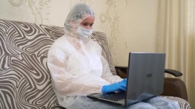 Hasta kadın koruyucu beyaz takım elbise giyiyor, tıbbi maske takıyor, mavi eldiven takıyor, evdeki dizüstü bilgisayarını kullanarak kanepeye oturuyor. Metin yazan, internette sörf yapan, uzaktan izole edilmiş hasta bir kız. Karantinaya alın. Salgın.